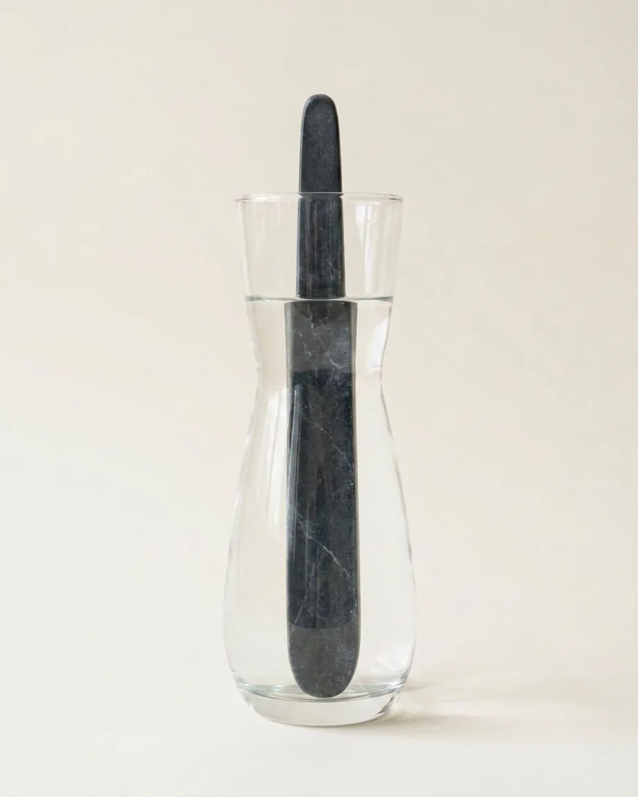 The Black marble wand standing in the glass carafe of water, against a plain pale background.